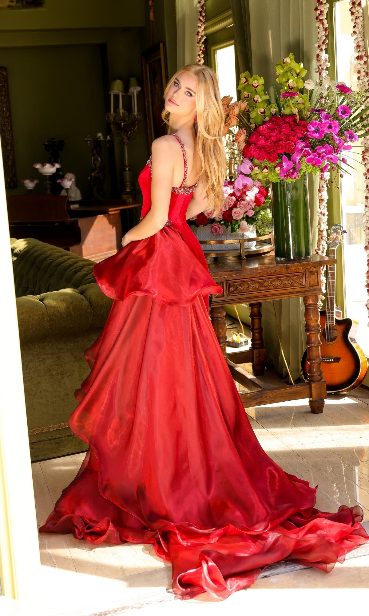 A woman in the Long Prom Dress 39556 by Ava Presley stands indoors, glancing over her shoulder, with a wooden table adorned with large floral arrangements and a guitar resting against it in the background.